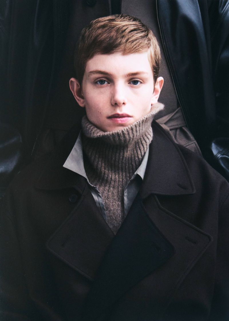Close-up portrait of a young person with short brown hair wearing a turtleneck, an open collared shirt, and a dark overcoat.