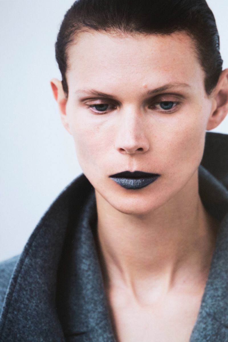 Headshot of a fair-skinned adult woman with dark, glossy lipstick, wearing a grey wool coat in a bright, neutral studio setting.