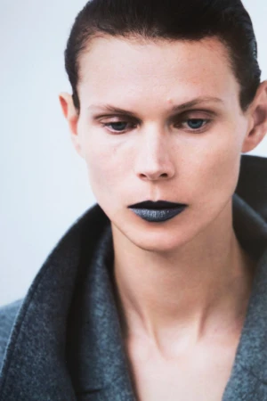 Headshot of a fair-skinned adult woman with dark, glossy lipstick, wearing a grey wool coat in a bright, neutral studio setting.