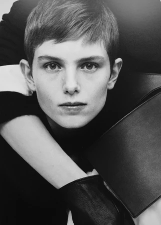 Close-up portrait of a person with short brown hair wearing a black sweater and leather gloves, holding a portfolio, black and white.