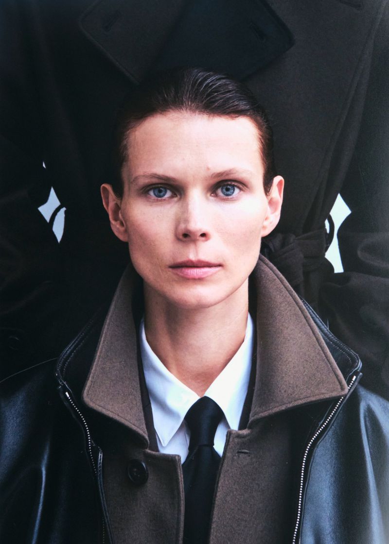 Fashion portrait of an adult woman in a black leather and wool coat, wearing a white button-down shirt with a black tie, against a dark backdrop.