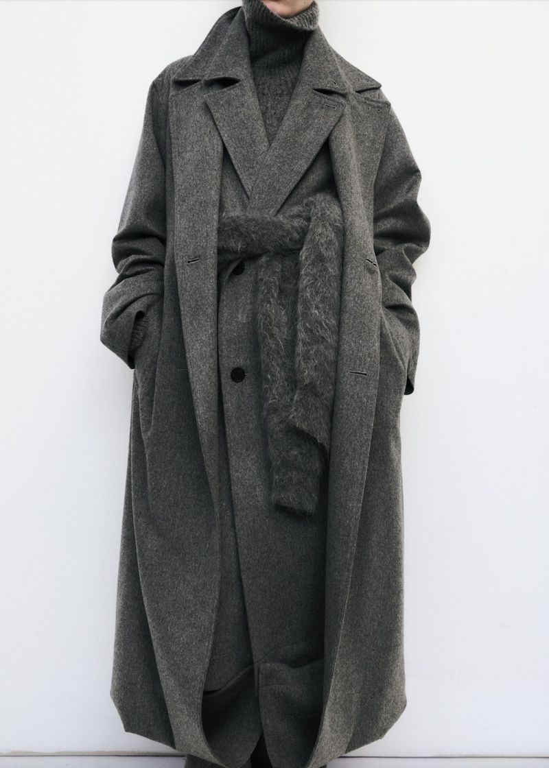 A gray, wool, belted overcoat on a fashion model stands in front of a white wall in a full shot. The model wears a matching turtleneck.
