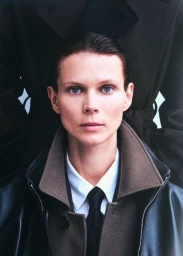Close-up portrait of a model wearing a collared shirt, black tie, and layered outerwear in Ssstein campaign