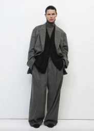 Model standing against a white seamless background wearing an oversized grey blazer, matching trousers, and a dark sweater.