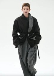 Model with short brown hair wearing a dark belted jacket and grey trousers against a clean white background