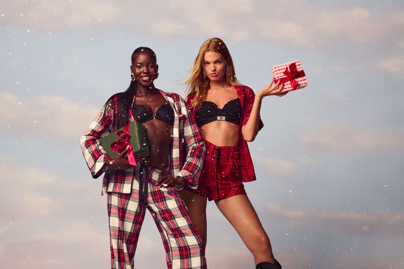Two women pose in plaid pajamas, holding gifts, against a snowy blue background, showcasing festive fashion apparel and holiday cheer.