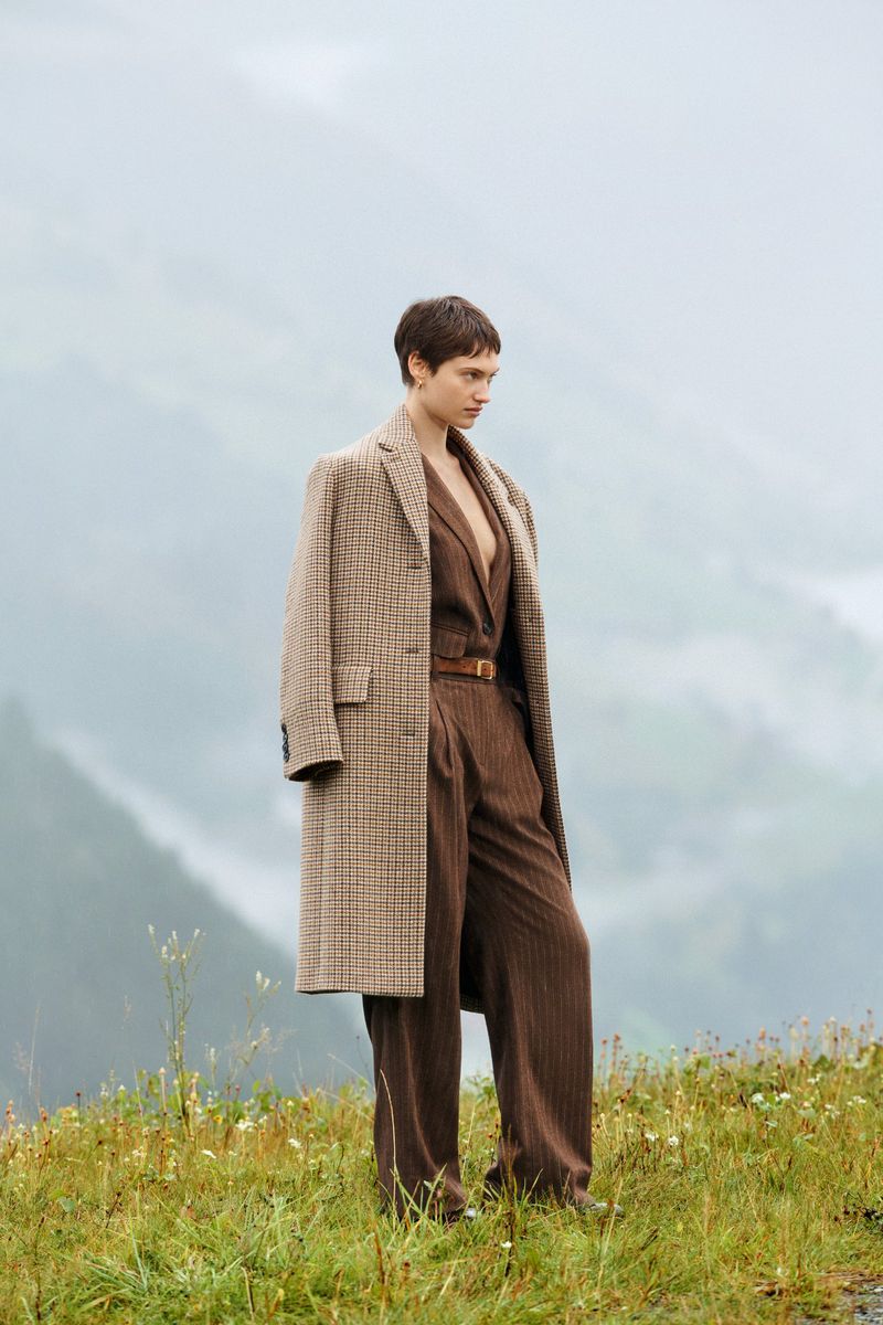 An adult female model is outdoors in a landscape wearing a brown pinstripe suit with a matching coat and belt, posing for a fashion editorial.