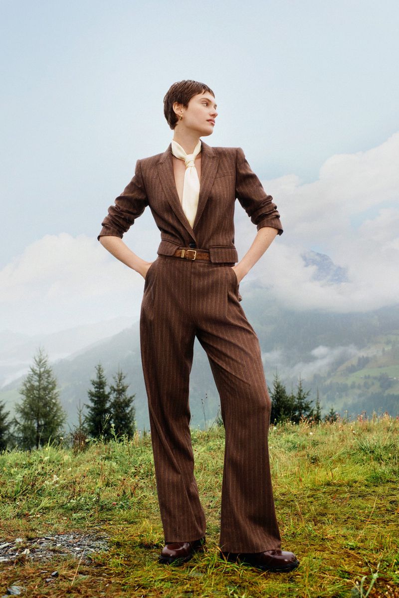 A young adult woman poses in a brown pinstripe suit with a white scarf, outdoors against a mountain backdrop under an overcast sky.