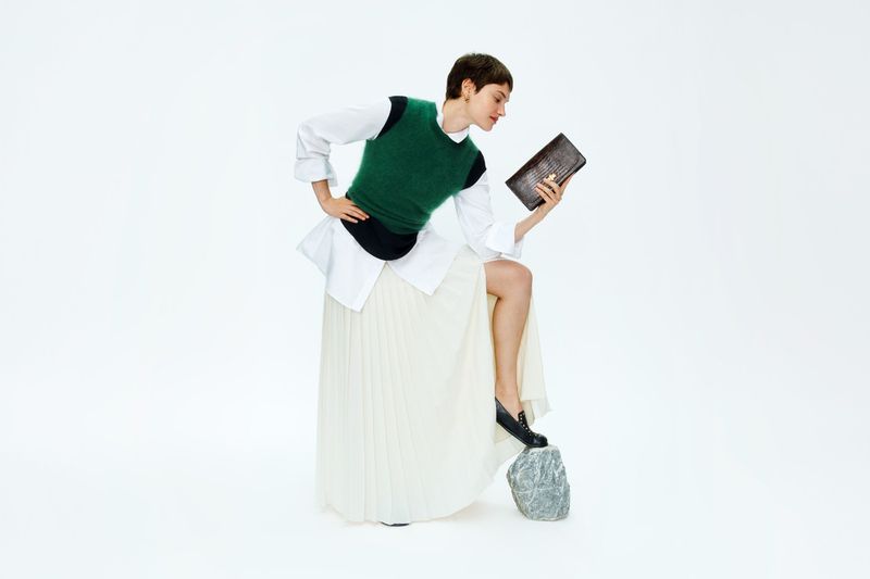 Woman in green sweater vest and long white skirt holds brown purse in a fashion pose on a white studio background.