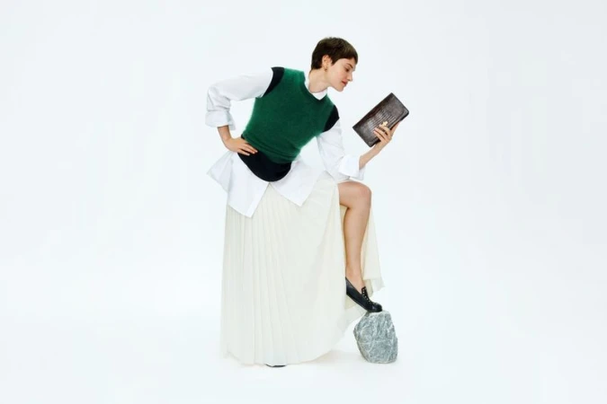 Woman in green sweater vest and long white skirt holds brown purse in a fashion pose on a white studio background.