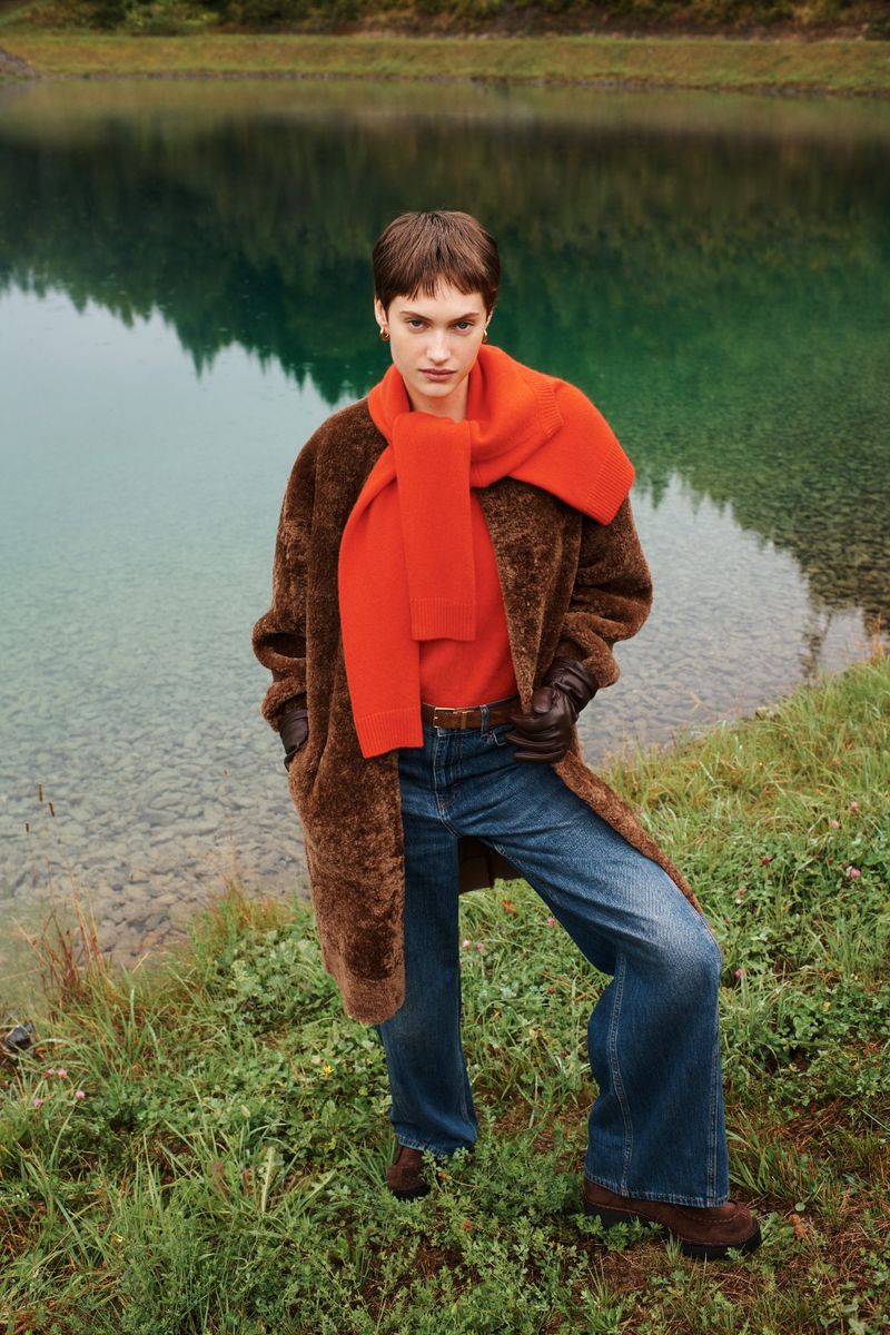 A young woman stands outdoors wearing a brown coat, orange scarf, and jeans, with a lake and forest in the background.