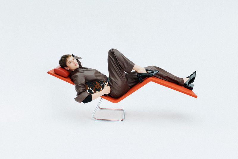 Woman in brown outfit reclines on orange chaise lounge in a fashion editorial shot against a white backdrop in a bright photo studio.