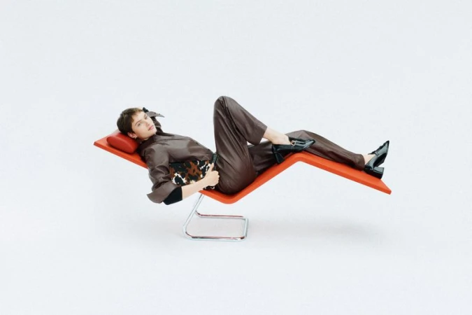 Woman in brown outfit reclines on orange chaise lounge in a fashion editorial shot against a white backdrop in a bright photo studio.