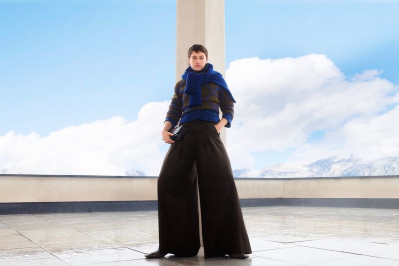 A woman in wide-legged pants, a blue scarf, and a sweater poses outdoors on a terrace with snowy mountains in the background.