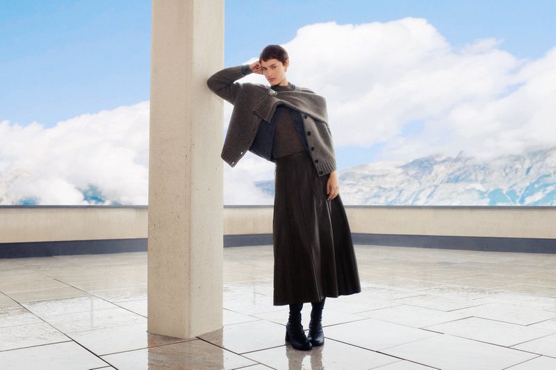 A fashionable woman poses in neutral tones against a scenic mountain backdrop, showcasing stylish knitwear and a flowing midi skirt.