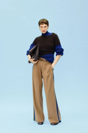 Fashionable woman in a brown, black, and blue sweater, tan slacks with blue stripes, carrying a black purse against light blue.