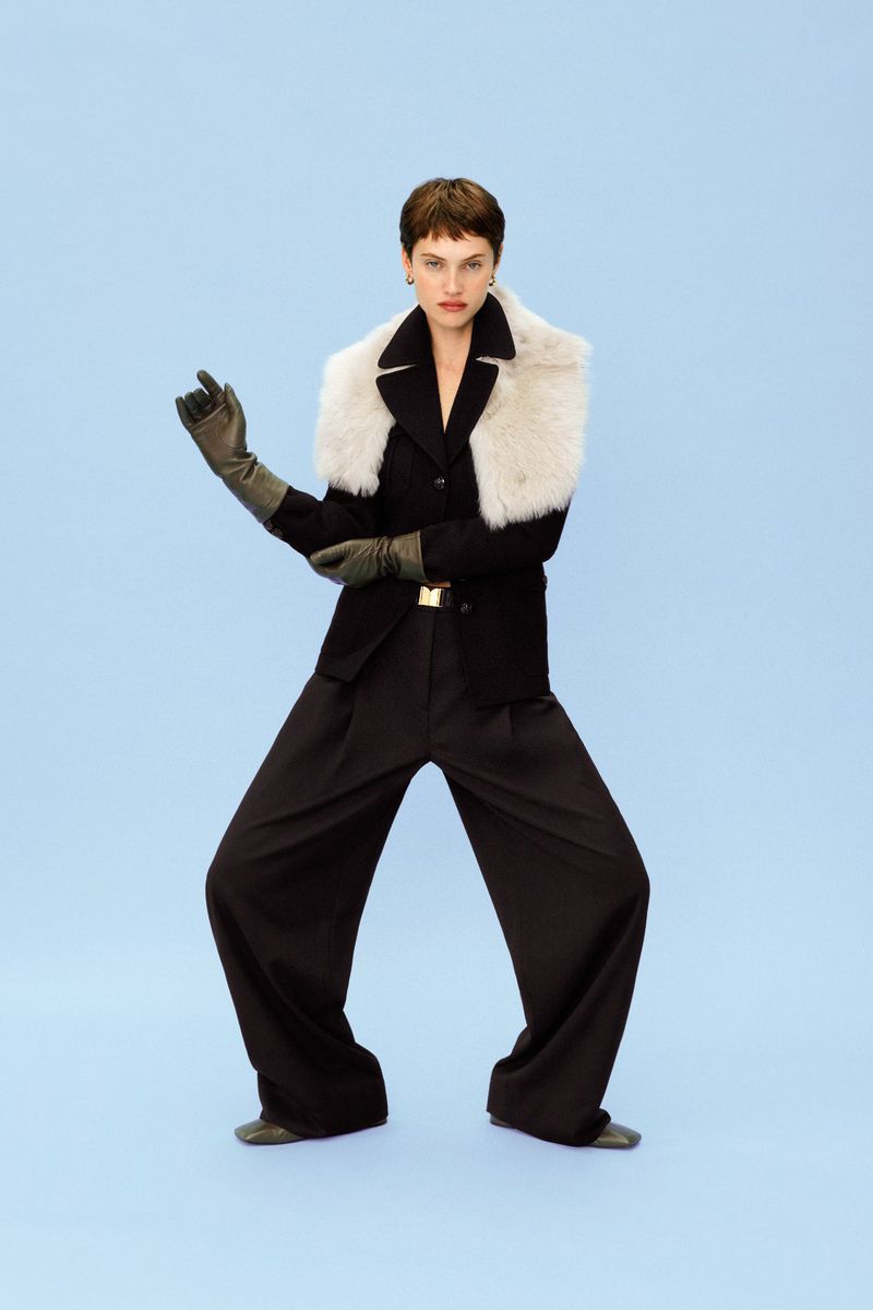 Elegant woman in a black blazer, wide-leg trousers, and gloves, accented by a white fur collar, posing against a serene sky-blue backdrop.
