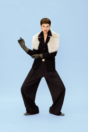 Elegant woman in a black blazer, wide-leg trousers, and gloves, accented by a white fur collar, posing against a serene sky-blue backdrop.