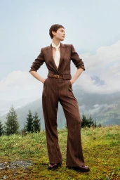 Model standing outdoors wearing a brown pinstripe suit and cream neck scarf against a mountainous background