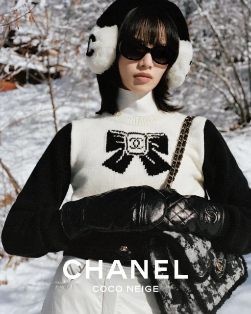 A woman in a Chanel sweater, earmuffs, and sunglasses poses in a snowy landscape, accessorized with gloves and a Chanel handbag.