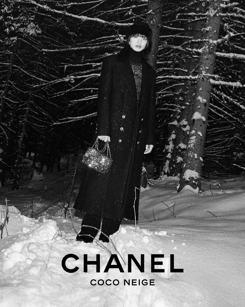 A woman in a coat stands in a snowy forest holding a purse in a black-and-white fashion photograph promoting Chanel's Coco Neige line.