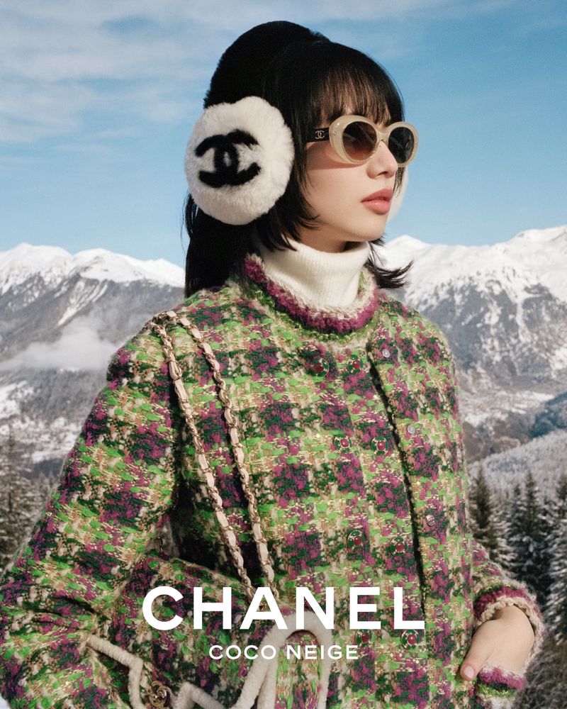 A woman models Chanel Coco Neige outerwear wearing earmuffs and sunglasses in a snowy mountain landscape on a sunny day.