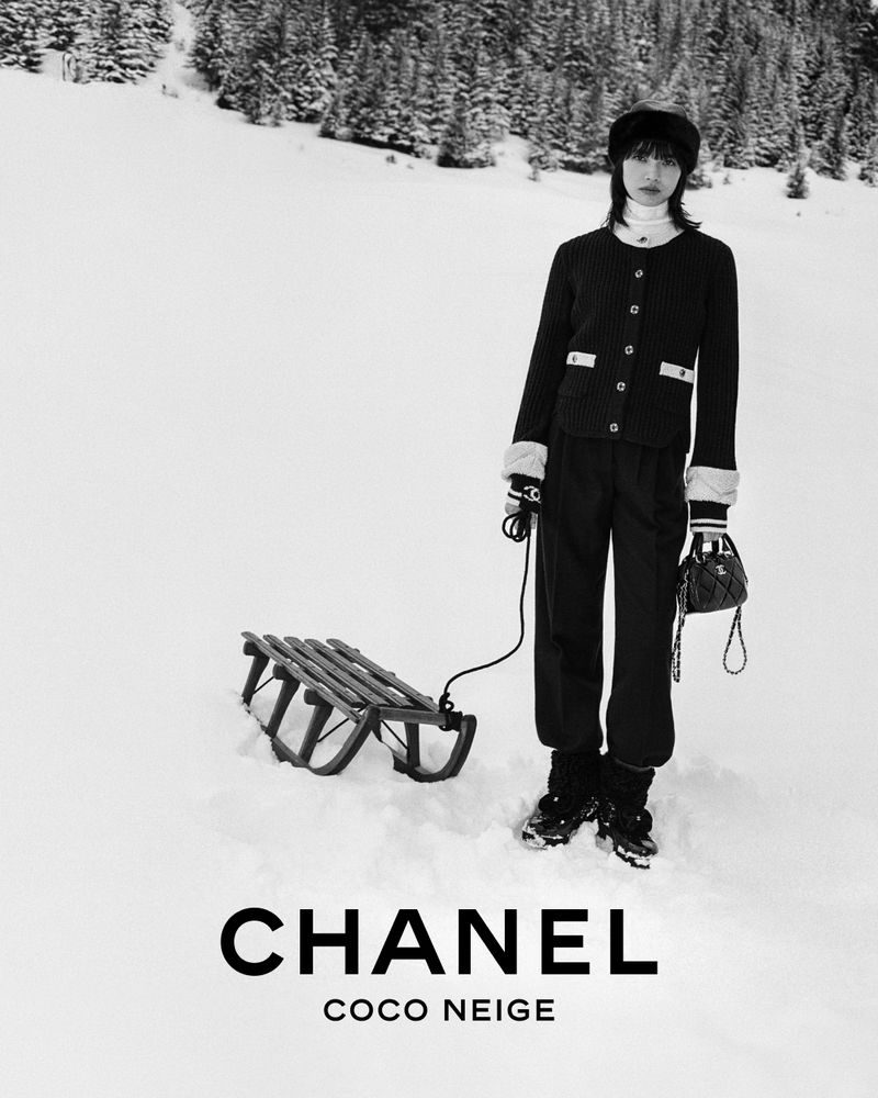 A woman wearing a black Chanel outfit stands in a snowy landscape pulling a sled, in a black and white editorial fashion shot.