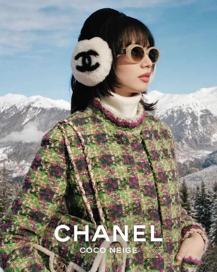 Nana Komatsu wearing white earmuffs with a Chanel logo and a green tweed jacket against a snowy mountain backdrop.