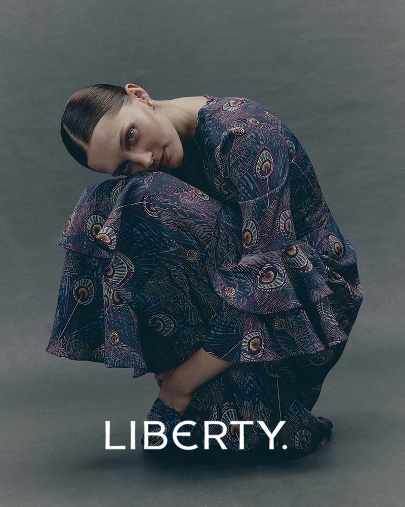 A fashionable young adult woman poses in a peacock feather print dress in a studio with neutral background lighting, showcasing a fashion editorial.
