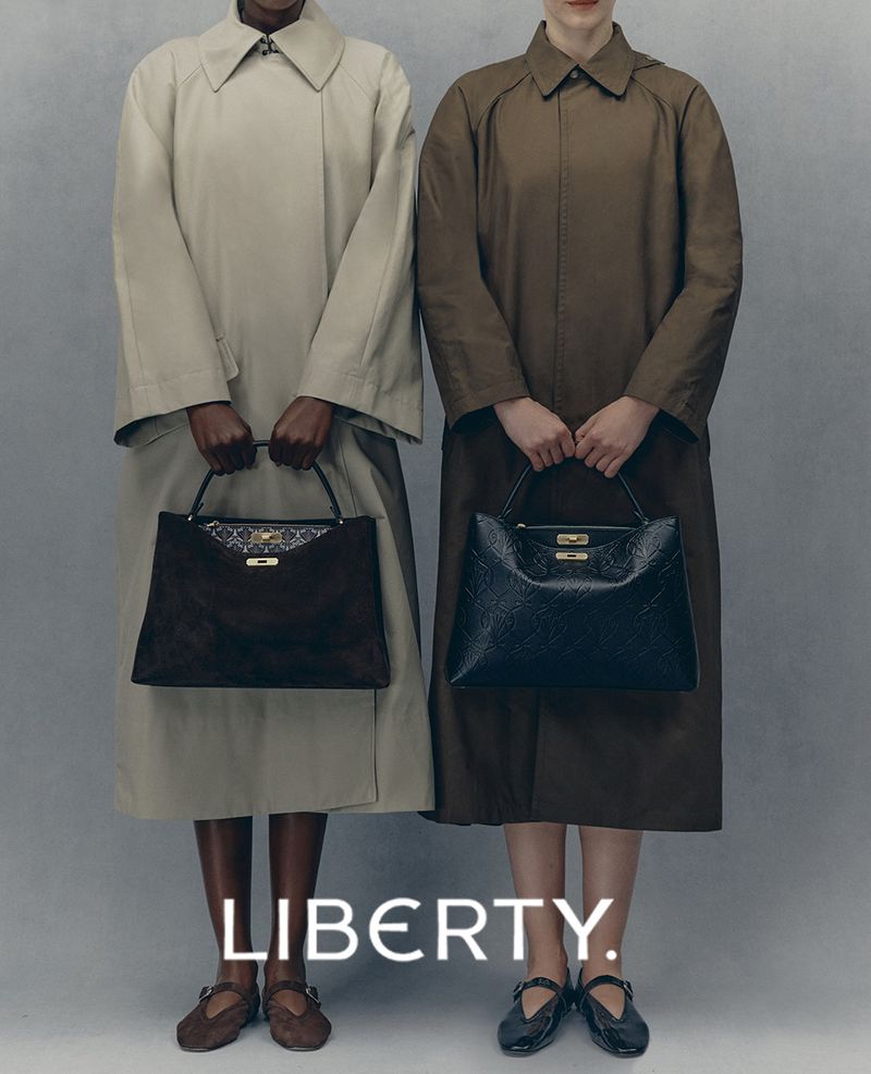Two women in coats holding matching designer handbags, standing in a photo studio against a neutral backdrop for a fashion editorial.