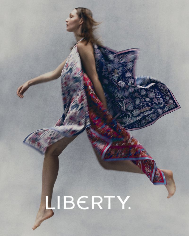 Fashion shot of a woman leaping with flowing floral print fabrics against a soft grey background, showcasing movement and style.