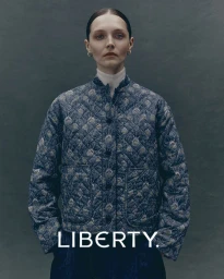 Model wearing a blue patterned quilted jacket against a dark grey studio background with Liberty branding at the bottom.
