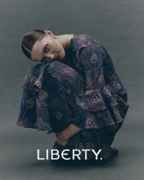 Model crouching wearing a long-sleeved patterned dress against a grey studio backdrop with the Liberty logo at the bottom.