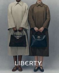 Two models wearing long coats and holding handbags against a grey studio backdrop for Liberty FW25 campaign.