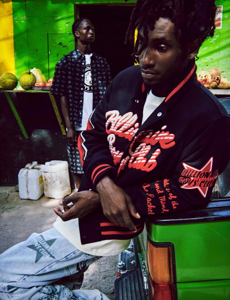 Two young Black men in streetwear pose in front of a green building with yellow accents. One wears a varsity jacket and jeans with logos.