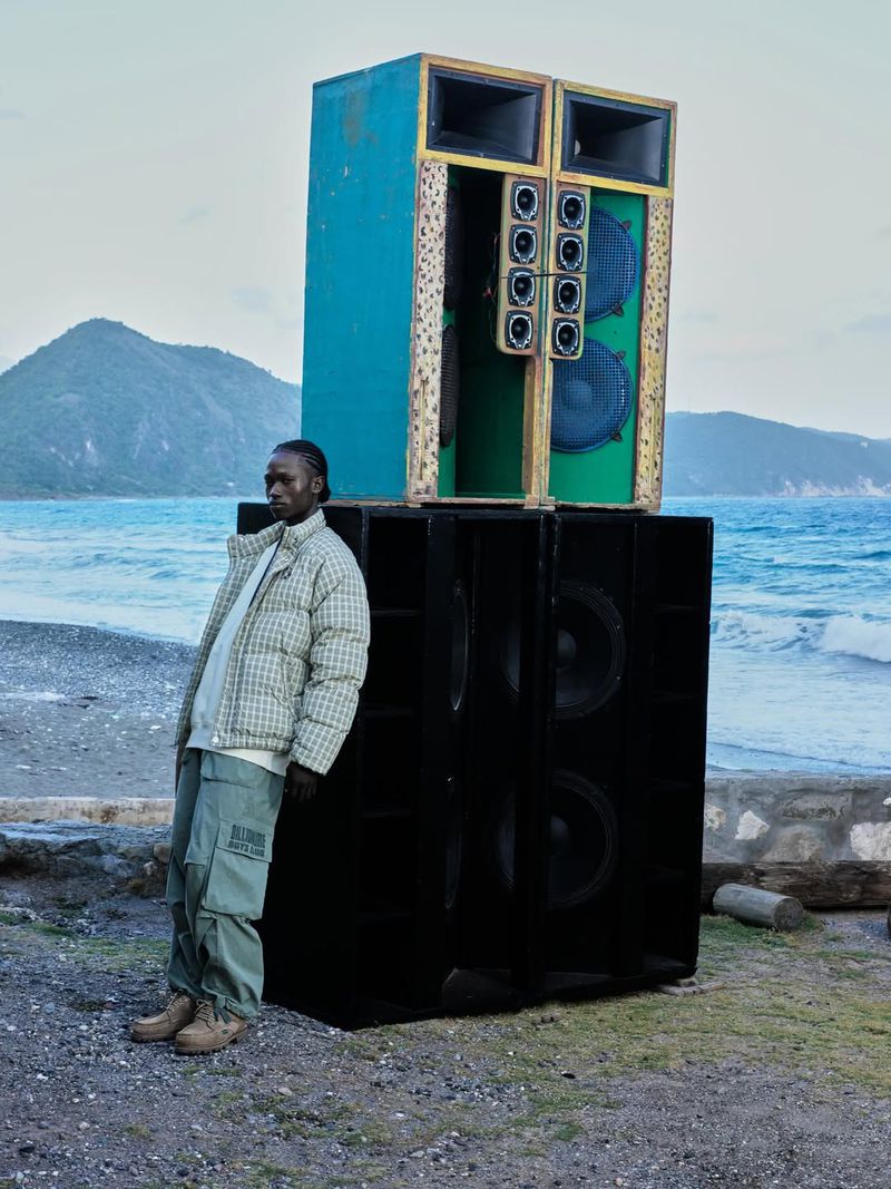 Man in patterned jacket leans against a large sound system on a rocky beach with the ocean and mountain in the background.