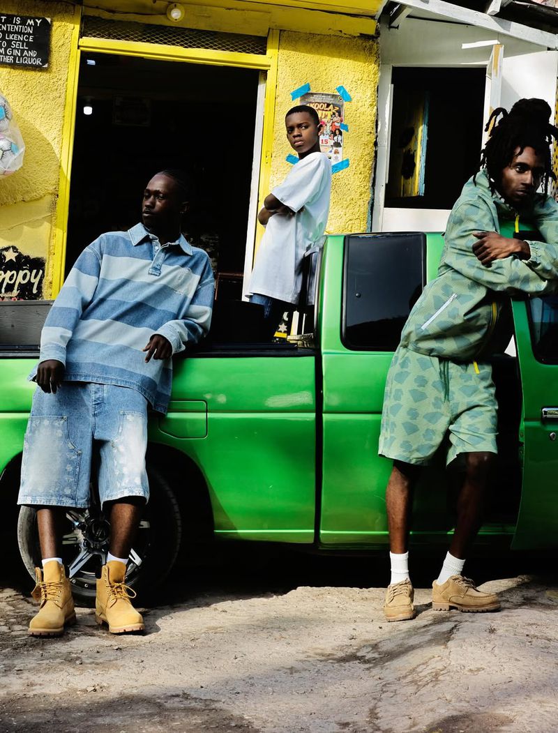 Three young men pose beside a green pickup truck in front of a yellow building. They wear casual clothing and boots outdoors.
