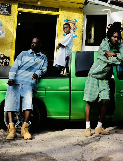 Three young men pose beside a green pickup truck in front of a yellow building. They wear casual clothing and boots outdoors.