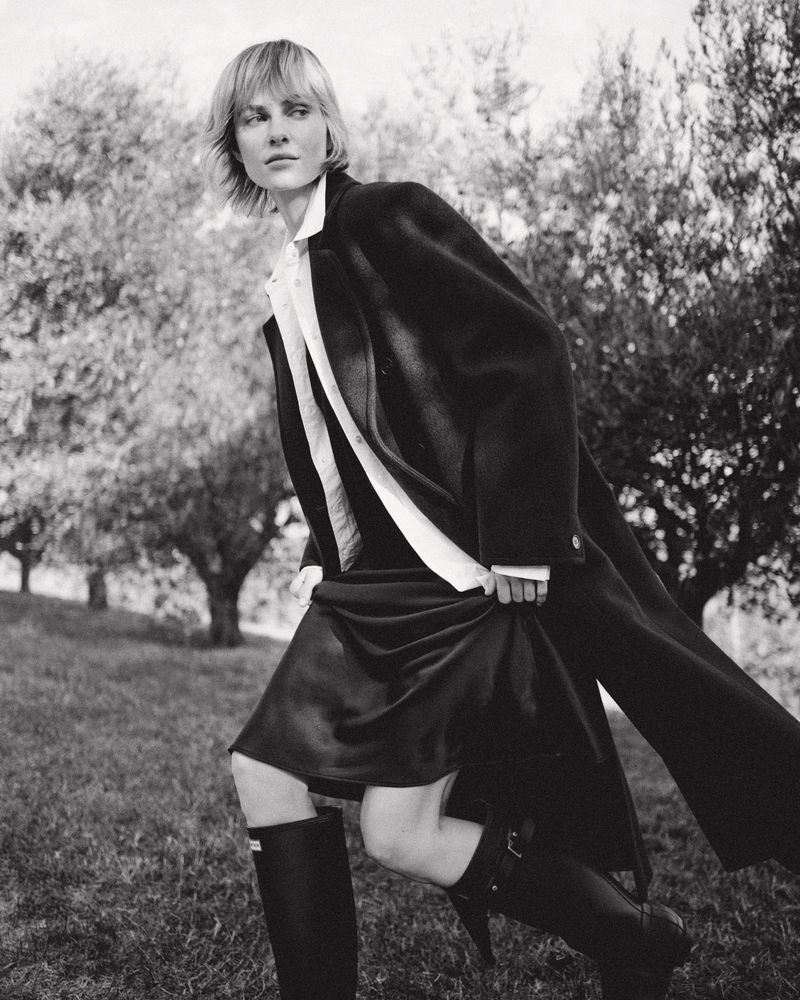 Monochrome shot of a fashionable adult woman in a coat, dress, and boots in a natural environment with trees in the background.