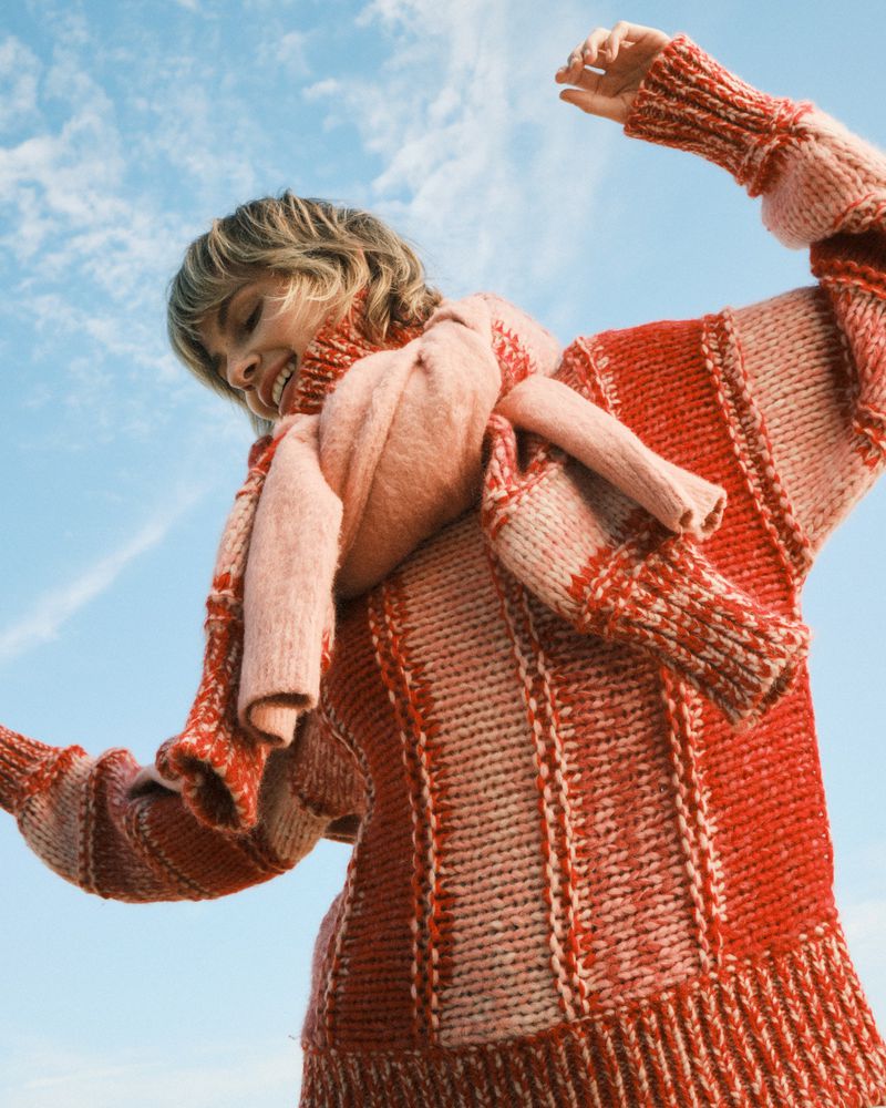 Woman in pink and red knit sweater and scarf stands against a blue sky, looking down and smiling, bathed in bright daylight.