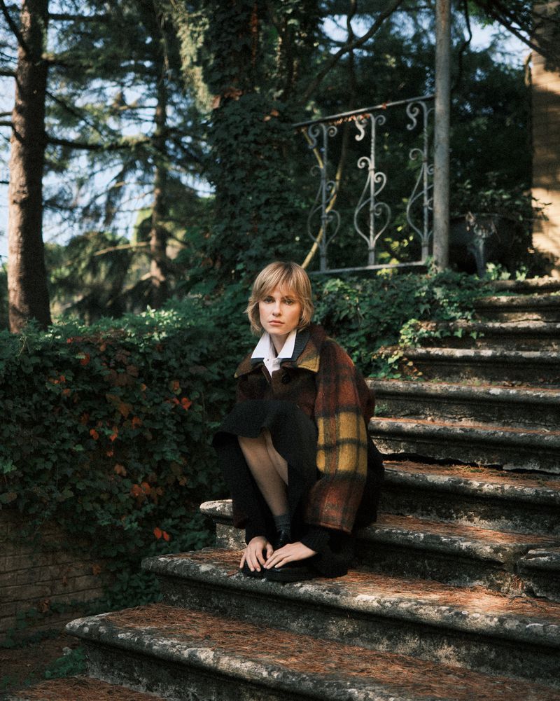 A young adult woman with short blonde hair in a patterned jacket and black skirt sits on stone steps outside in a wooded area.