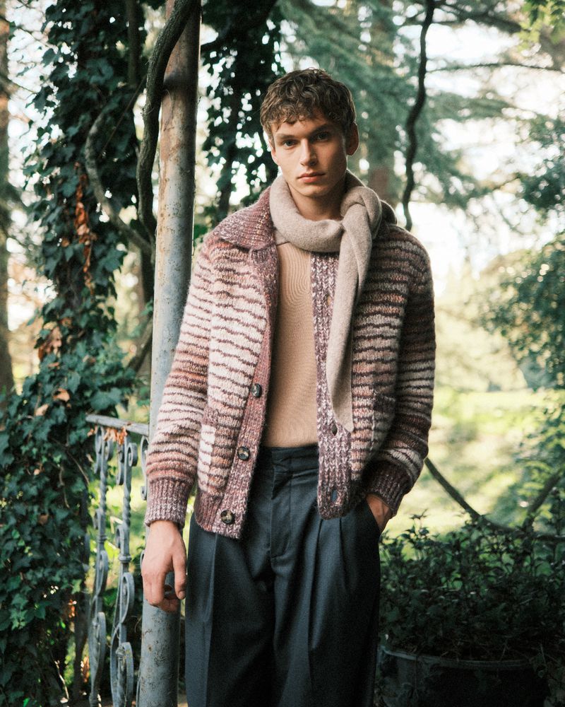 Adult man in a knitted cardigan and scarf poses outdoors in a natural setting with trees and foliage, styled in neutral and earthy tones.