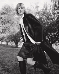 Model in dark coat and tall boots walking through an outdoor grassy landscape with trees under natural light