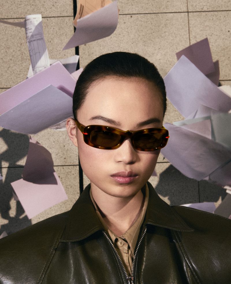 Portrait of a woman wearing tortoiseshell sunglasses and a green leather jacket, with white paper in the background. Beauty, fashion.