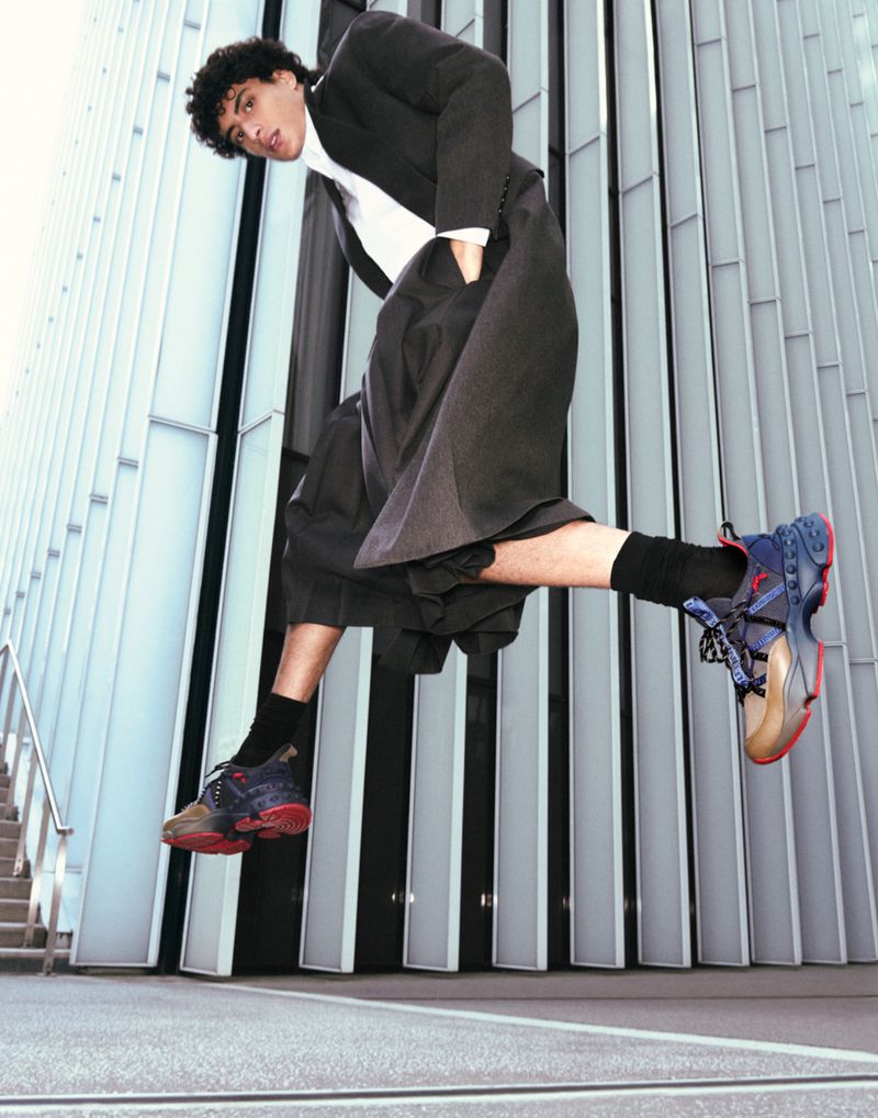 Man in black suit jacket and wide-legged trousers leaping mid-air against a modern building backdrop, showcasing unique style.