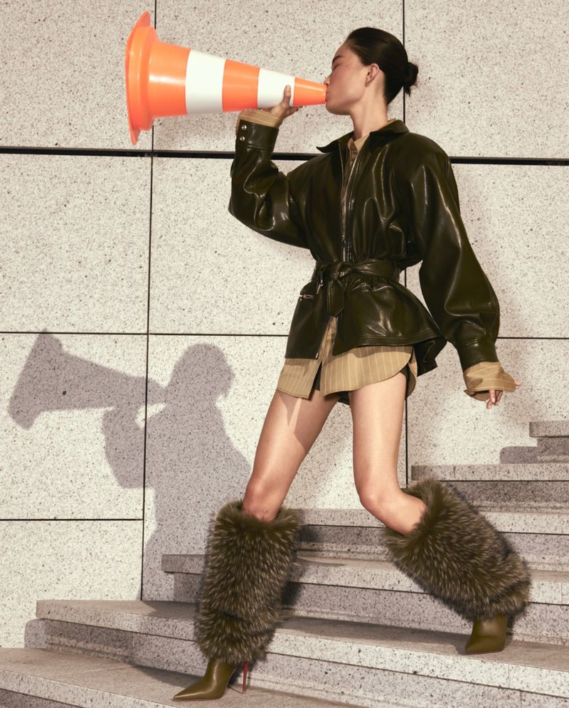 A woman in a stylish outfit is standing on stairs, holding an orange traffic cone to her mouth like a megaphone, fashion editorial shot.