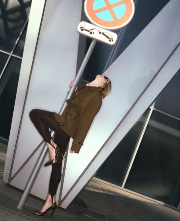 Fashionable woman in brown jacket and leggings poses with a no-parking sign in an urban setting, wearing gold heels and sunglasses.