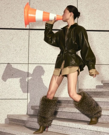 Model in olive green leather jacket and furry boots holding a traffic cone on stairs for Christian Louboutin Spring 26 campaign.
