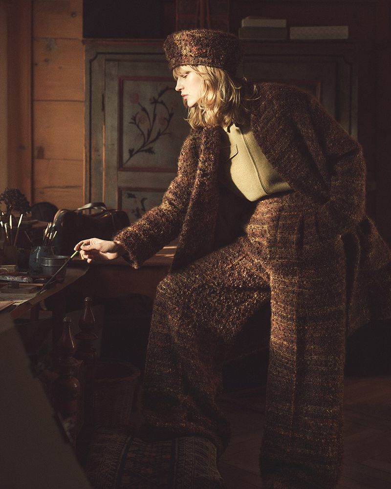 Woman in a brown tweed suit and hat is painting at a table with art supplies and brushes. Soft lighting illuminates the scene.