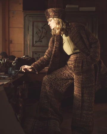Woman in a brown tweed suit and hat is painting at a table with art supplies and brushes. Soft lighting illuminates the scene.